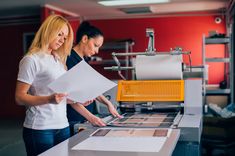 two women standing in front of a large screen printing something on it's surface