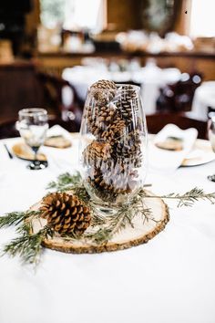 pine cones are placed in a glass vase on a table with white linens and place settings