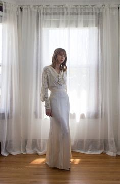 a woman standing in front of a window wearing a long white dress with lace sleeves