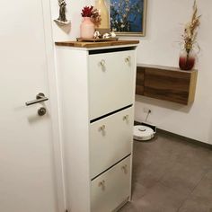 a white cabinet with three drawers in a bathroom next to a toilet and flower vase