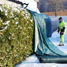 a man standing next to a green fence covered in snow and covering it with a tarp