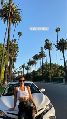 a woman sitting on the hood of a white sports car in front of palm trees