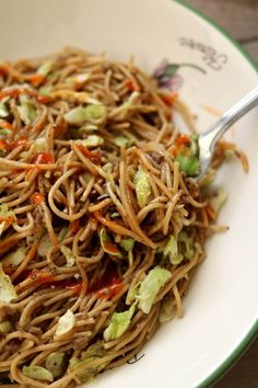 a white bowl filled with noodles and vegetables on top of a wooden table next to a fork