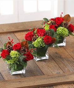 three clear vases filled with red roses and greenery on top of a wooden table