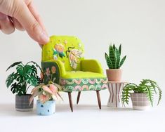 a hand is pointing at a miniature green chair next to small potted plants and houseplants