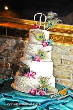 a three tiered cake with peacock feathers and flowers on the top is displayed in front of a fireplace