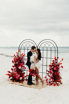 a man and woman kissing in front of an iron gate with flowers on the beach