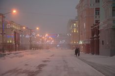 a man walking down a snow covered street in the middle of winter with lights on