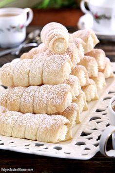 powdered sugar covered pastries are stacked on a white plate next to cups and saucers