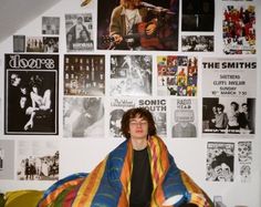 a young man sitting on top of a bed under a blanket covered in pictures and posters