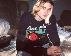 a young man sitting on top of a bed with his hand to his face and looking at the camera