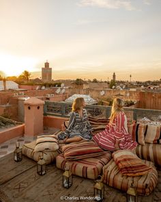 two women sitting on top of a wooden floor next to pillows and lamps in front of a cityscape