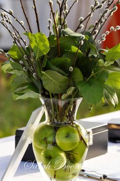 a vase filled with lots of green apples on top of a table next to flowers