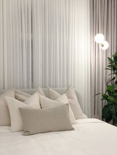 a white bed topped with lots of pillows next to a window covered in sheer curtains