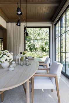 a dining room table with white chairs and vases on the table in front of large windows