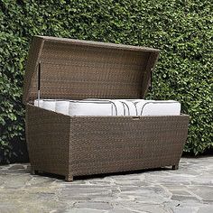 a brown wicker storage box sitting on top of a stone floor next to a green bush