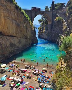 people are on the beach in front of an old stone bridge and blue water under it