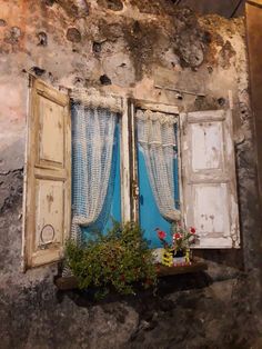 an open window with curtains and flowers in front of it on the side of a building