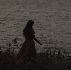 a woman in a long dress is standing by the water at night with her arms outstretched