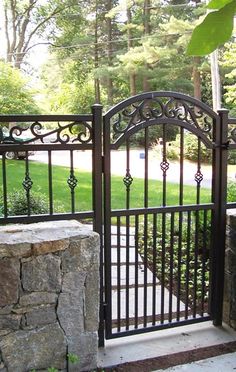 an iron gate in front of a stone wall and green yard with trees behind it