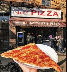 a person holding up a slice of pizza in front of a restaurant on the street