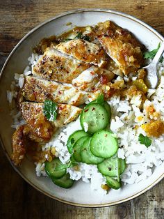 a white plate topped with rice and chicken next to cucumbers on top of a wooden table