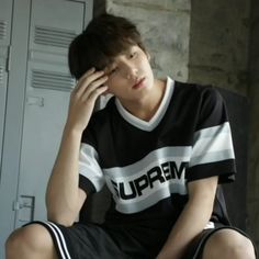 a young man sitting in front of lockers holding his hand to his face while wearing a black and white t - shirt with the word supreme on it