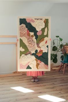 a woman holding up a large piece of art with flowers on the wall behind her