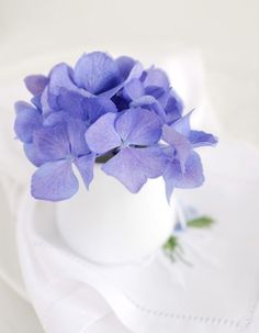 a white vase filled with purple flowers sitting on top of a white cloth covered table