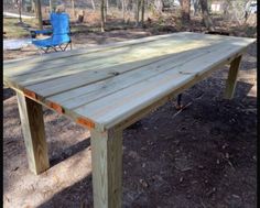 a wooden table sitting in the middle of a forest next to a blue lawn chair