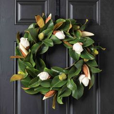 a wreath with white flowers and green leaves hanging on a black front door, decorated with greenery