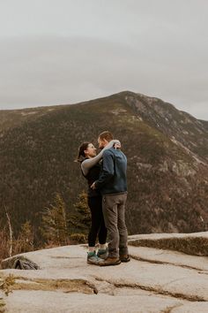 a man and woman standing on top of a mountain hugging each other with mountains in the background