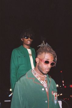 two people standing next to each other in front of a dark background wearing sunglasses and green clothing