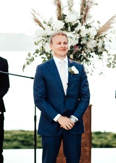 a man in a blue suit standing next to a flower filled ceremony arch with white flowers