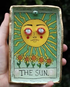 a hand holding up a small ceramic sun with red eyes and the words, the sun