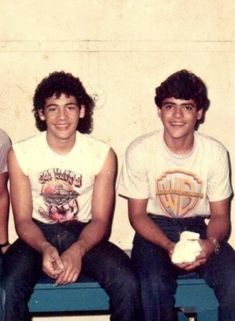 three young men sitting next to each other in front of a white wall and blue bench