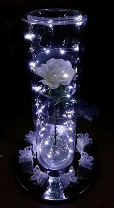 a clear vase with white flowers in it on a black plate and some lights around it