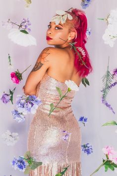 a woman with red hair wearing a gold dress and flowers around her neck, standing in front of a white backdrop