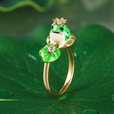 This whimsical piece captures the essence of nature's beauty and adds a touch of regal charm to your ensemble. Made from sterling silver and hand painted with enamel, this enchanting ring features an adorable frog wearing a crown, poised gracefully amidst a lotus leaf. The intricate flawless enamel work coupled with white stones, make this an eye-catching piece. This exquisite combination of elements evokes a sense of tranquility and harmony. It will surely become a conversation starter and a cherished addition to your jewelry collection.Carat Weight: 0.545 ctStone Size: 2.5,3.5 mmStone Type: Jeulia® StoneNumber of Stones: 3 Stone Shape: RoundStone Color: Diamond WhiteWeight: 5.8 gWidth: 2.2 mmHeight: 10.4 mmThickness: 1.3 mmMaterial: 925 SilverPlating Color: Yellow Gold Wearing A Crown, Frog Jewelry, 9th Anniversary, Frog Prince, Lotus Leaves, White Stones, Lotus Leaf, White Stone, Anniversary Sale