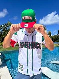 a man wearing a mexico baseball jersey and hat standing in front of a swimming pool