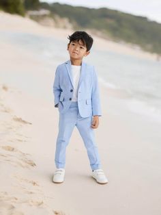 a little boy standing on top of a sandy beach next to the ocean wearing a blue suit