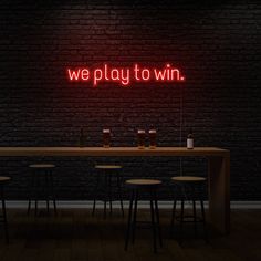 a neon sign that says we play to win on the wall above a table with four stools