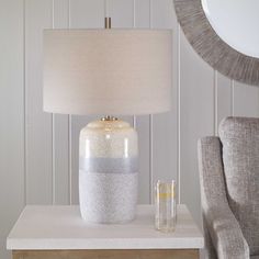 a table with a lamp and a glass on it next to a chair in front of a white wall