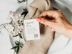 a person holding up a business card on top of a white plate next to flowers