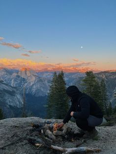 a man sitting on top of a mountain next to a campfire with mountains in the background