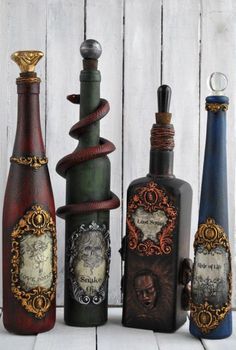 five different colored bottles are lined up against a white wooden wall, one has an ornate design on the top