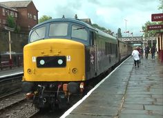 a yellow and black train on the tracks at a station with people walking near by