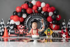 a baby sitting in front of a birthday cake with balloons and race cars around it