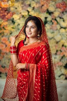 a woman in a red and gold sari posing for the camera with flowers behind her