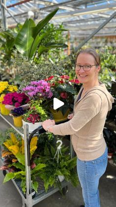 a woman standing next to a cart full of flowers in a greenhouse filled with potted plants